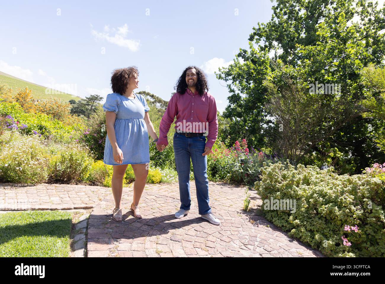 Paare stehen auf gemauertem Gartenweg und halten Hände unter sonnigem Himmel mit Sträuchern Stockfoto