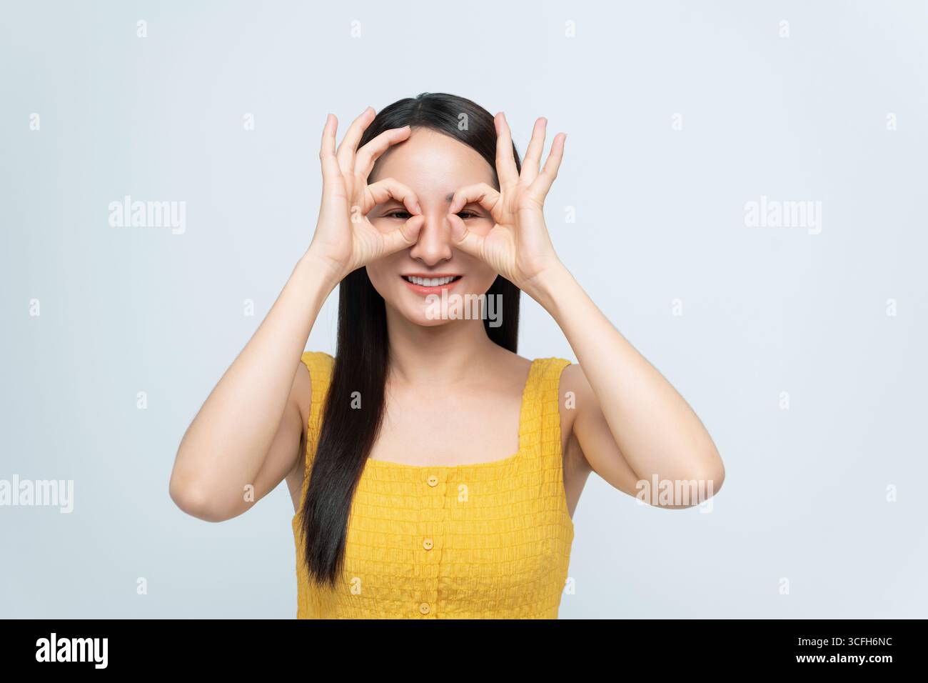Lächelnde junge Frau in einem gelben Kleid, die eine spielerische Handbewegung mit den Fingern über den Augen macht, wie eine Brille. Stockfoto