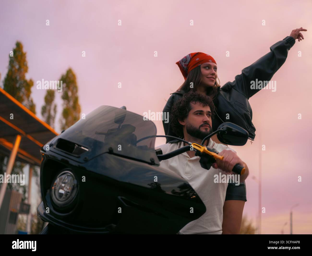 Ein junges Bikerpaar auf schwarzem Motorrad an einer Tankstelle. Frau, die auf dem Beifahrersitz steht, den Arm gestreckt hat und zeigt. Dynamische Szene, Freiheit, Stockfoto Ein junges Bikerpaar auf schwarzem Motorrad an einer Tankstelle. Frau, die auf dem Beifahrersitz steht, den Arm gestreckt hat und zeigt. Dynamische Szene, Freiheit, Stockfoto