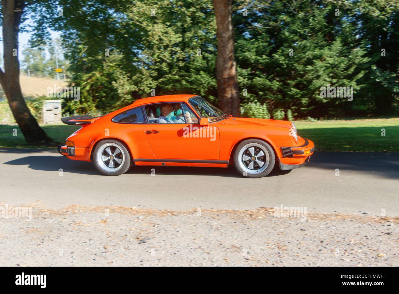 Klassischer oranger Sportwagen Porsche 911, der auf einer von Bäumen gesäumten Straße in einem sonnigen Park mit grünem Laub und blauem Himmel fährt. Stockfoto