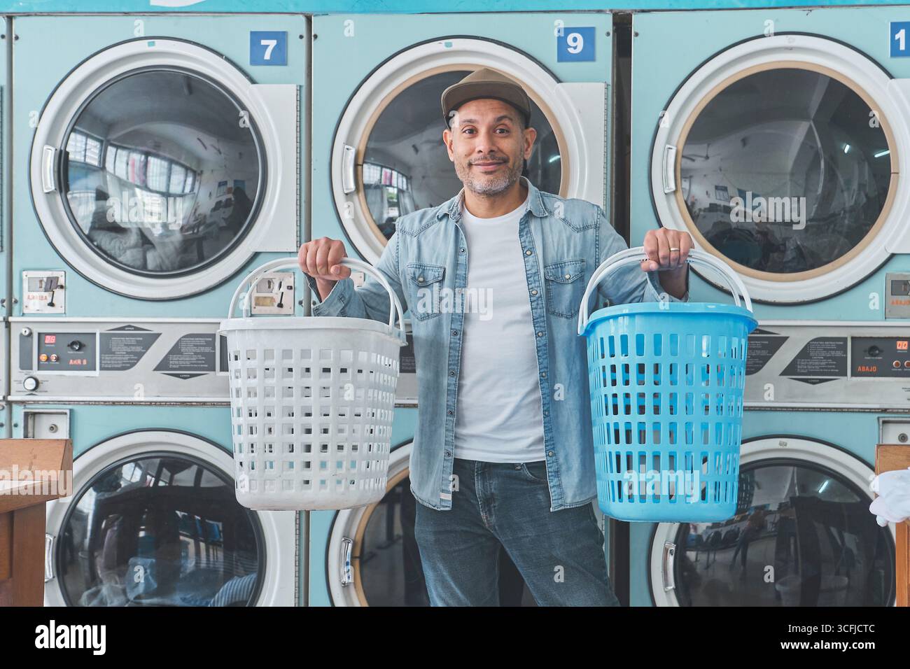 Kunde hält zwei Wäschekörbe, während er Selbstbedienungs-Waschmaschinen in einem Waschsalon benutzt Stockfoto