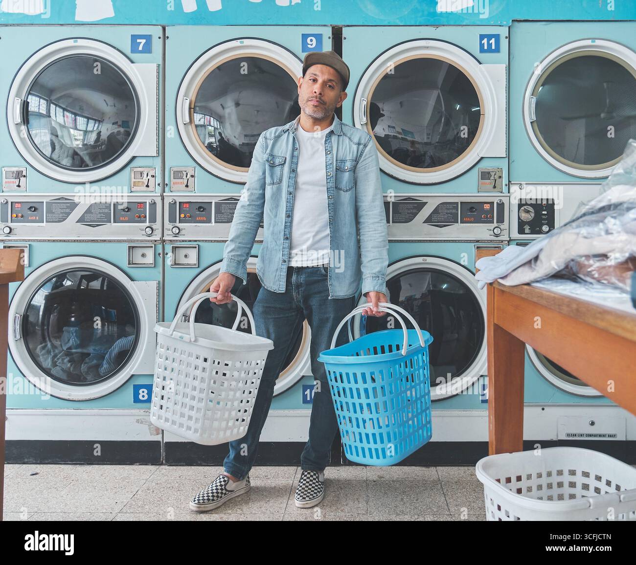 Der junge Mann hält zwei Wäschekörbe, lächelnd und blickt weg, in einem Waschsalon voller Waschmaschinen Stockfoto