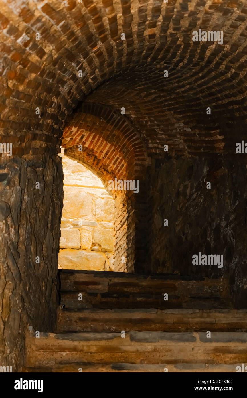 Warmes Licht strahlt aus einem Bogengang oben auf einer Steintreppe aus, mit strukturierten Ziegelwänden, die eine einladende Atmosphäre in einem alten Gebäude schaffen. Stockfoto