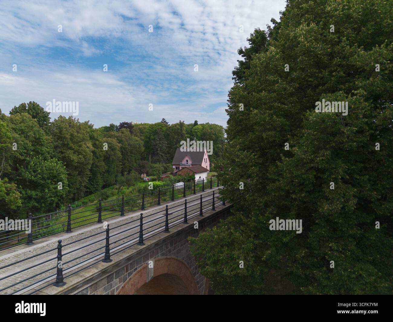 Wunderschöner Blick auf eine alte Eisenbahnbrücke, umgeben von üppigen grünen Bäumen, mit einem bezaubernden rosa Haus im Hintergrund vor einem blauen Himmel Stockfoto