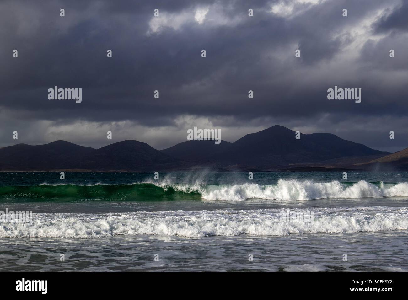 Dramatische Brandung mit grünen Wellen, die unter dunklen Sturmwolken auf das Ufer stürzen und Berge in der Ferne aufsteigen Stockfoto