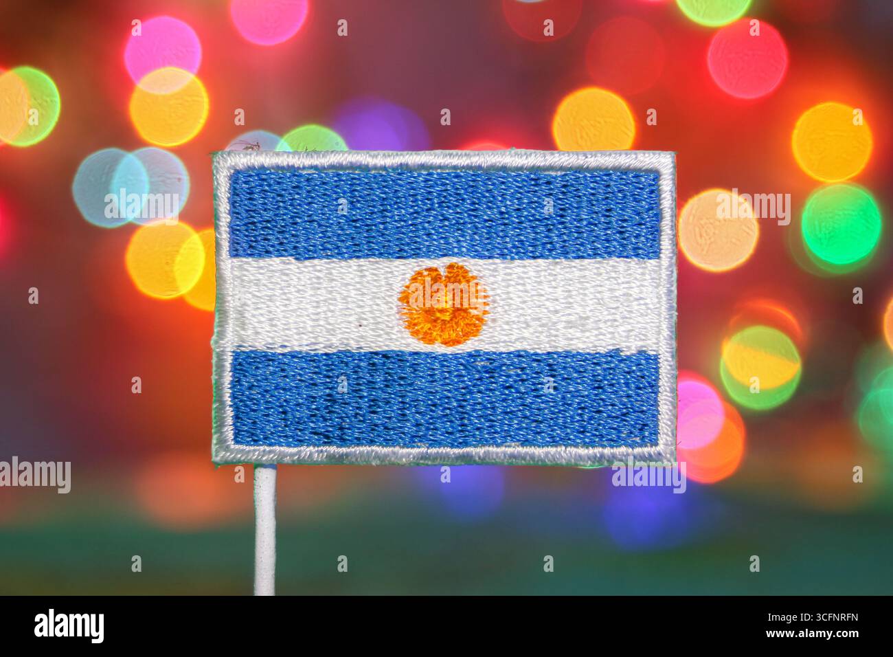 Argentinische Flagge mit goldener „Sonne des Mai“-Stickerei vor leuchtenden Bokeh-Lichtern, die an ein Feuerwerk und nationale Festlichkeiten erinnern. Stockfoto