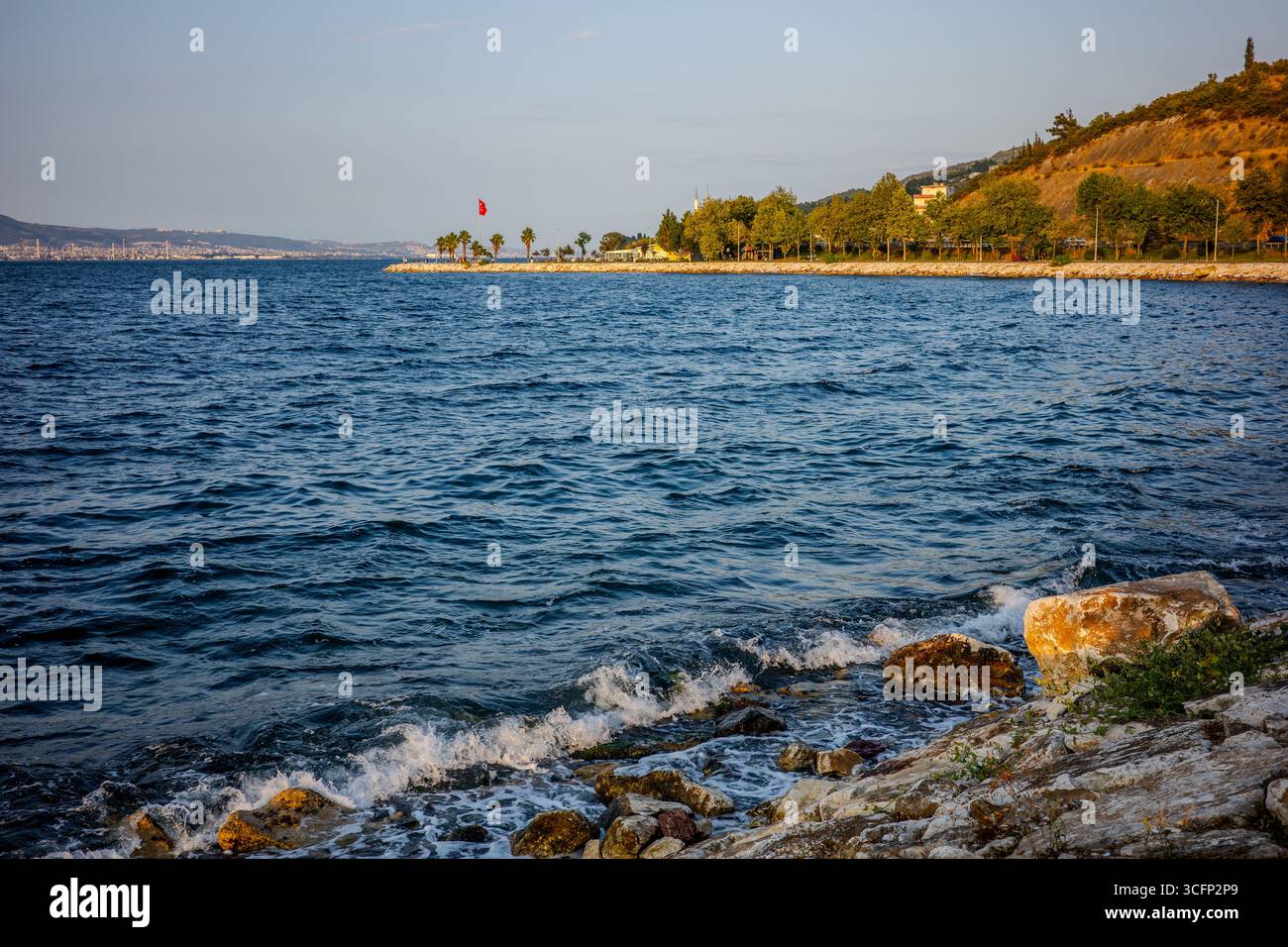 Wellen brechen gegen die felsige Küste des Marmarameers bei Karamürsel, Kocaeli, Türkiye, mit einer von Bäumen gesäumten Promenade und einem Hügel im Hinterland Stockfoto