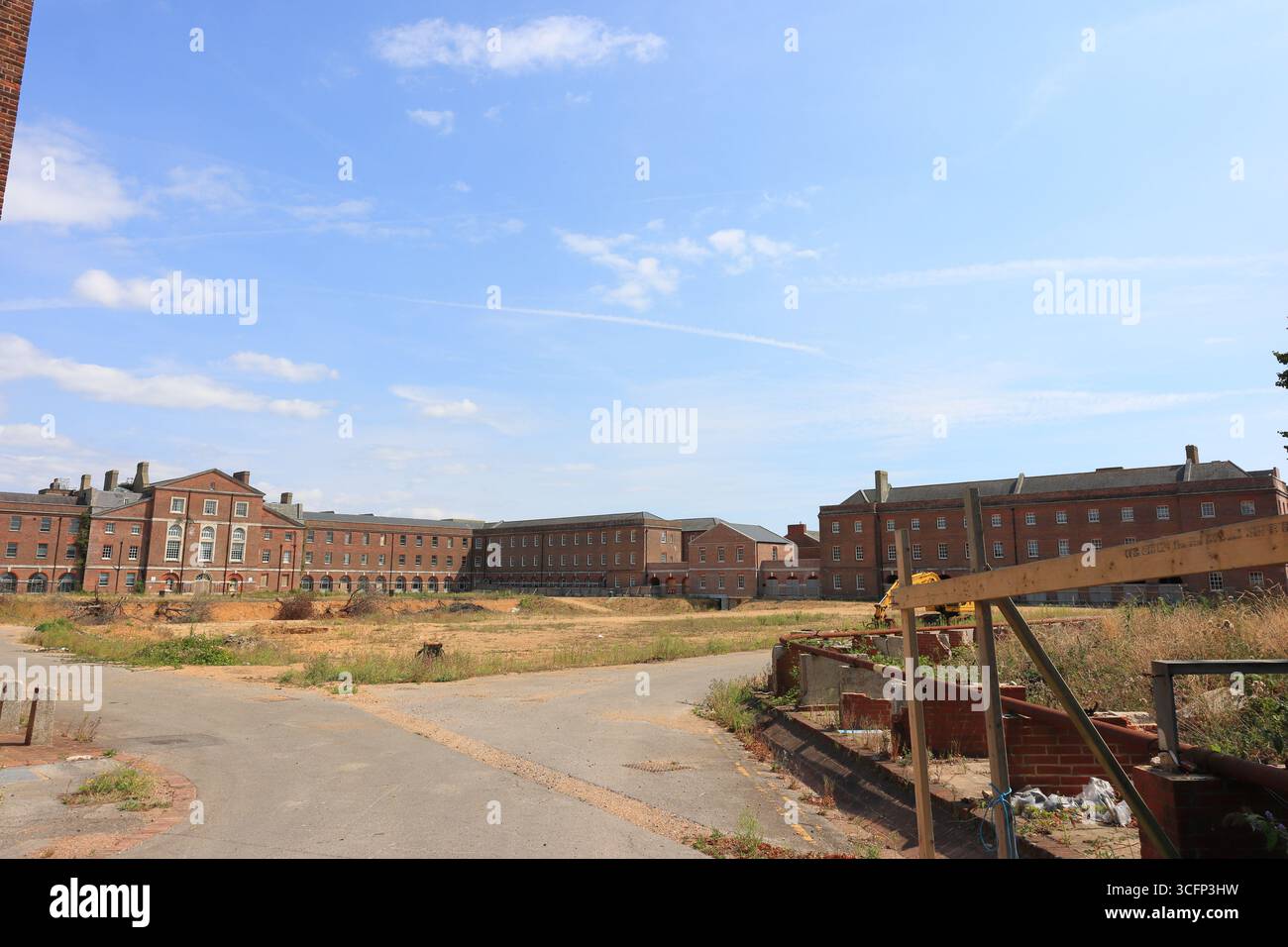 Auf dem Gelände des ehemaligen Krankenhauses Haslar wird ein weites Freigelände von großen, roten Backsteingebäuden mit Schornsteinen und Fensterreihen flankiert – wahrscheinlich ehemalige Stationen oder Verwaltungsblöcke. Baumaterialien liegen verstreut im Vordergrund und deuten auf eine aktive Sanierung hin. Die Mischung aus grasbewachsenen und gepflasterten Oberflächen unter einem hellen Himmel erinnert an einen Moment des architektonischen Wandels. Illustratives, redaktionelles Bild, Besuch des ehemaligen Krankenhauses Haslar in Gosport, Hampshire, England. August 2025. Stockfoto