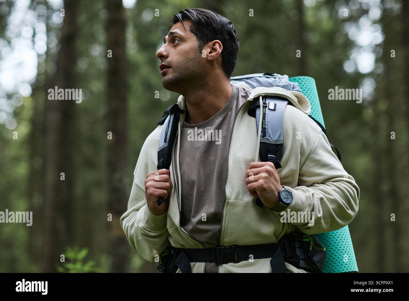 Junger Erwachsener kaukasischer Mann, der mit Rucksack durch den Wald wandert, mit fokussiertem Ausdruck aufblickt, die Natur während des Outdoor-Abenteuers erforscht, lässige Kleidung trägt, kurze dunkle Haare Stockfoto