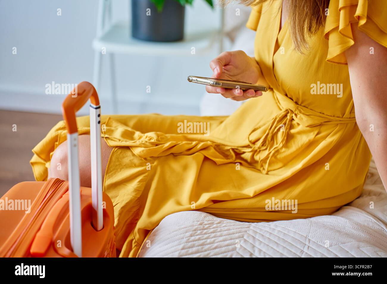Frau in gelbem Kleid, die auf dem Bett in der Nähe des orangefarbenen Koffers sitzt und das Smartphone benutzt und darauf wartet, das Haus zu verlassen. Weibliche Reisende, die Informationen im Mobi überprüft Stockfoto