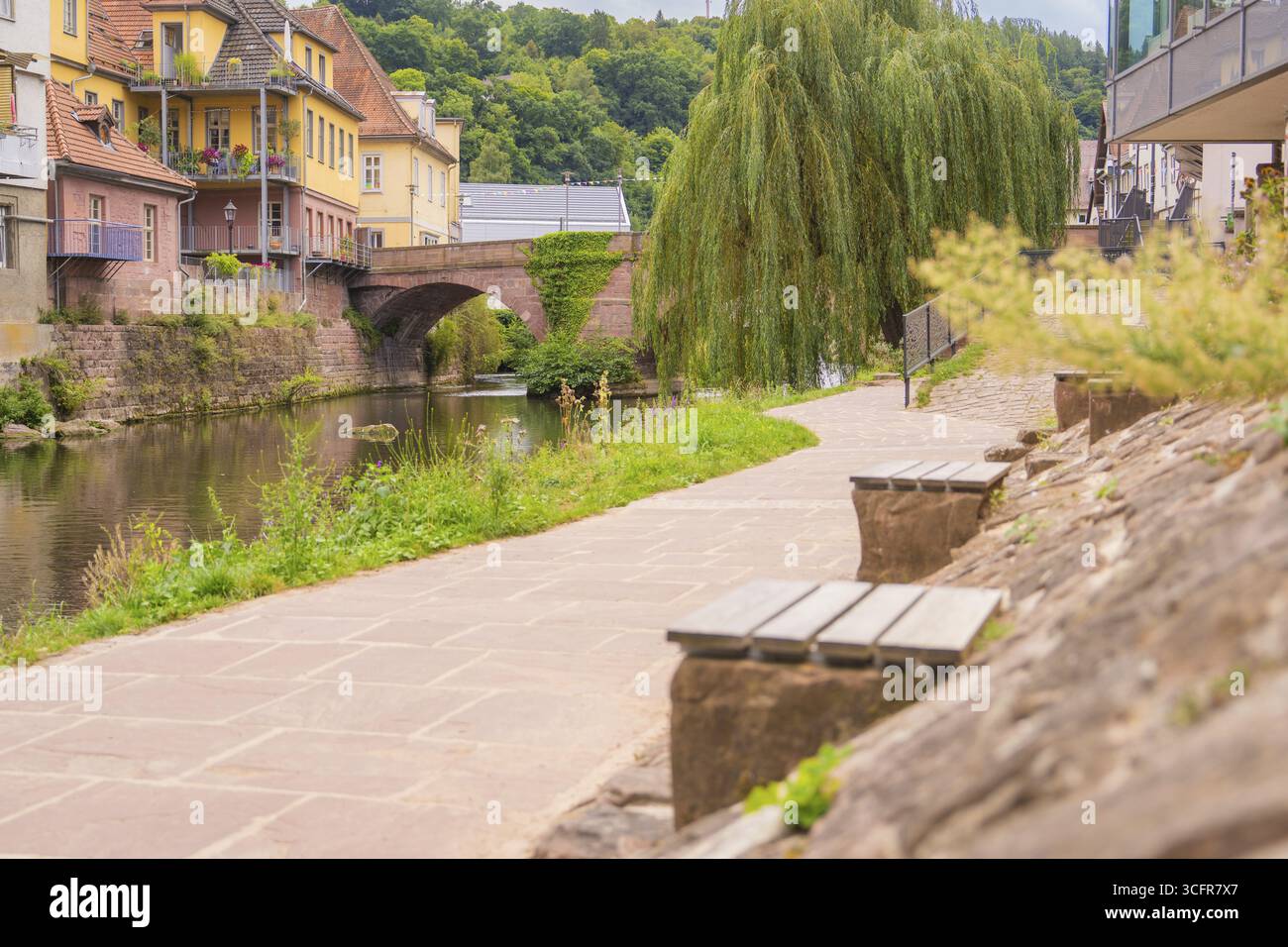 Gepflasterter Weg entlang eines Flusses mit Steinmauer und Bänken, eine Brücke im Hintergrund, kleine Stadt Perle Calw, Schwarzwald, Deutschland Stockfoto