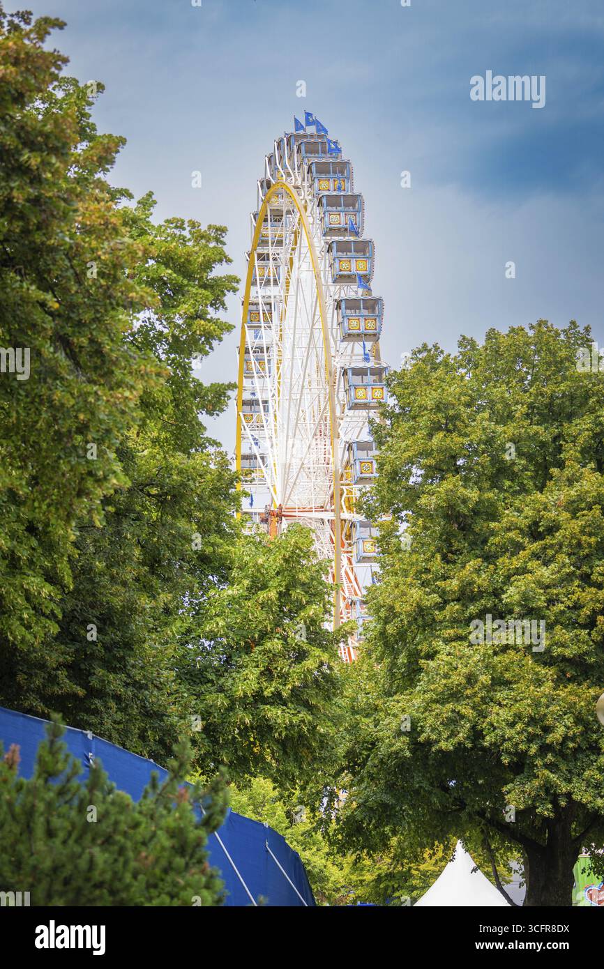 Riesenrad versteckt hinter dichten grünen Bäumen unter blauem Himmel, Olympiapark, München, Deutschland Stockfoto
