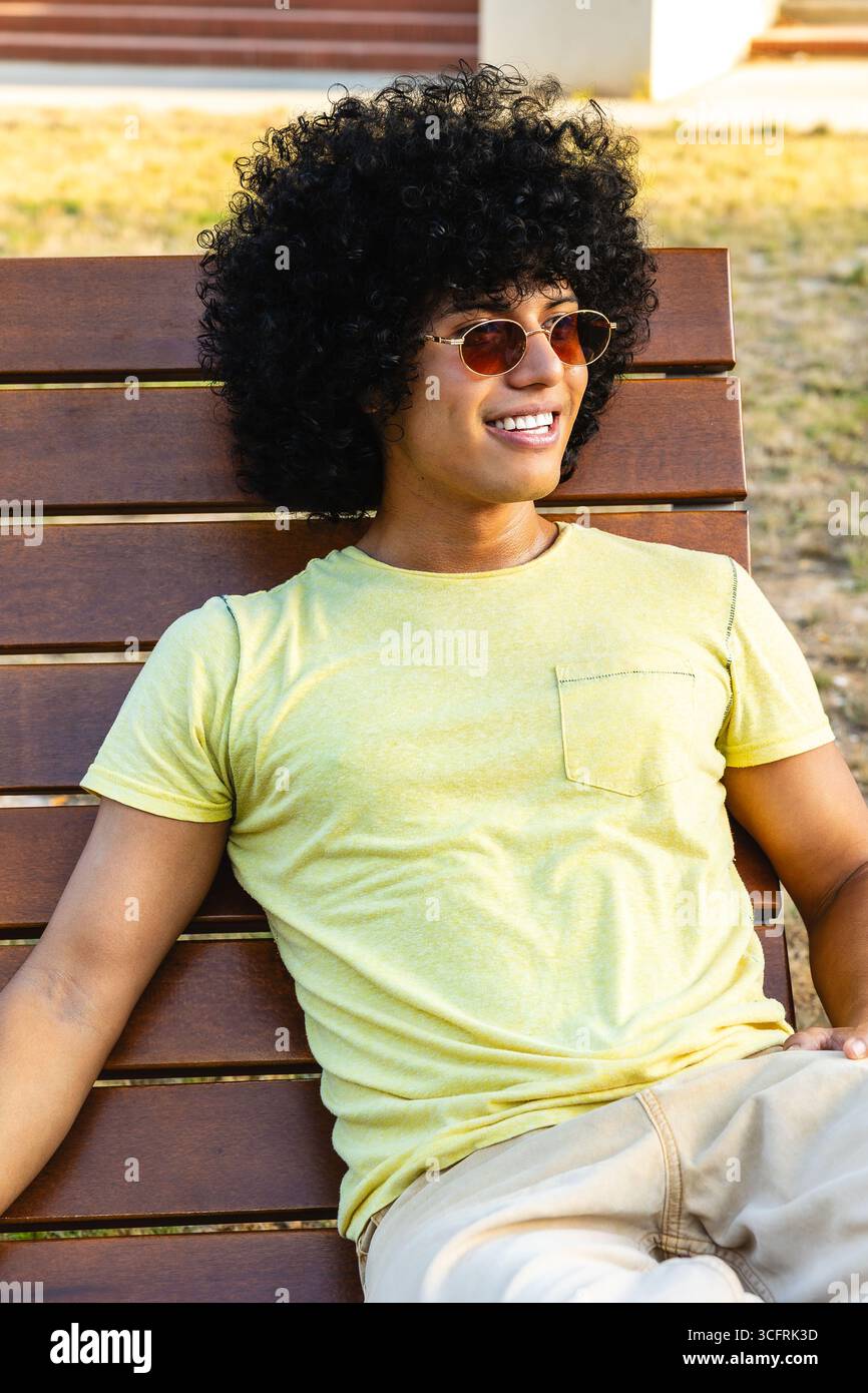Junger Mann mit Afro-Frisur, der sich auf der Bank im Freien entspannt Stockfoto