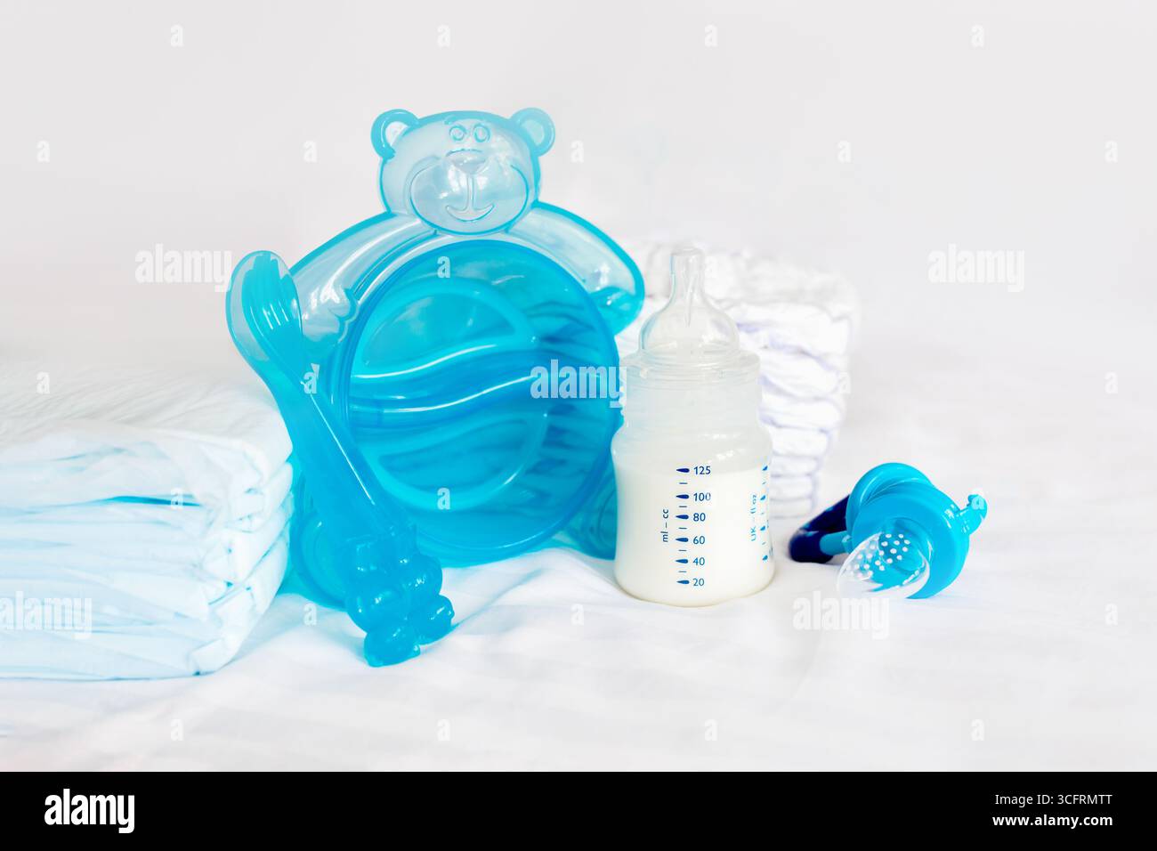 Babygeschirr-Set. Milchflasche, Fütterungsknabber, blaues Besteck, Löffel, Gabel, Windeln, Einwegwindeln auf dem Hintergrund eines weißen Bettes Stockfoto