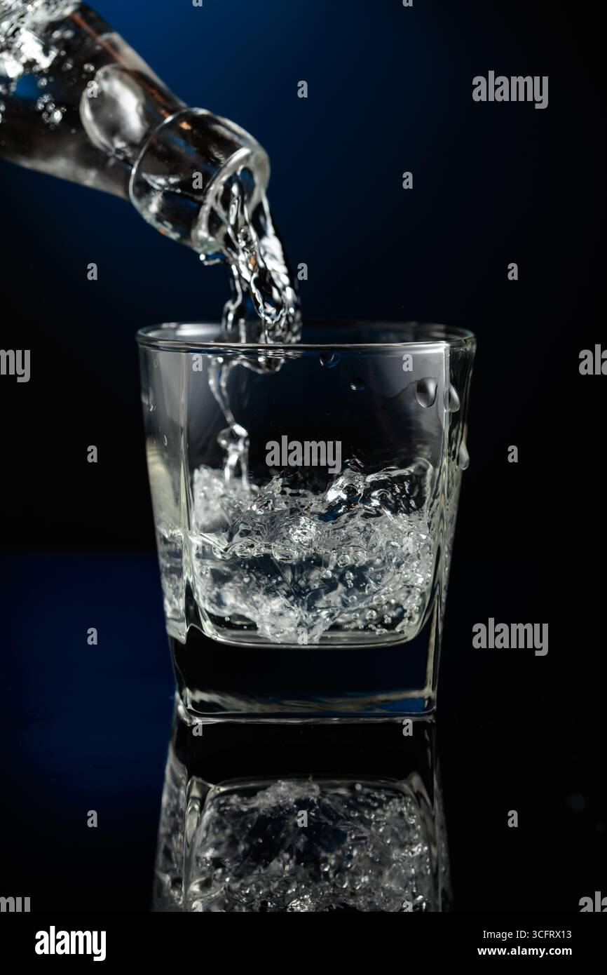 Kristallklarer Wodka oder Gin wird über Eis in ein Glas gegossen. Wodka mit Eis auf schwarzem reflektierendem Hintergrund. Stockfoto