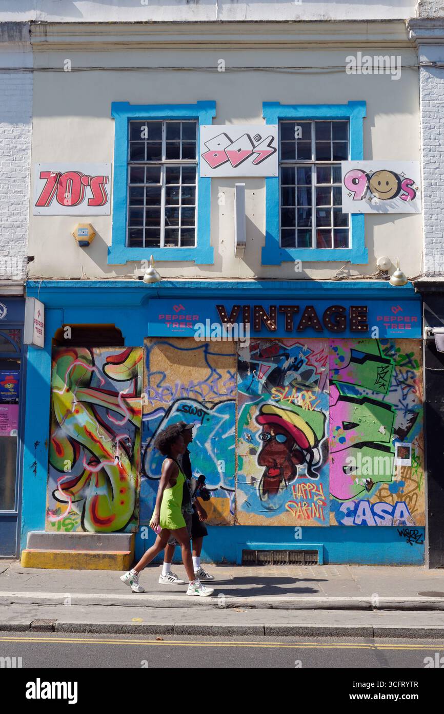 Ein Paar läuft an einem bewachten Schaufenster mit Graffiti vorbei, mit Frauen in gelbem Outfit in der Gegend von Notting Hill, London. England, 25. August 2025 Stockfoto