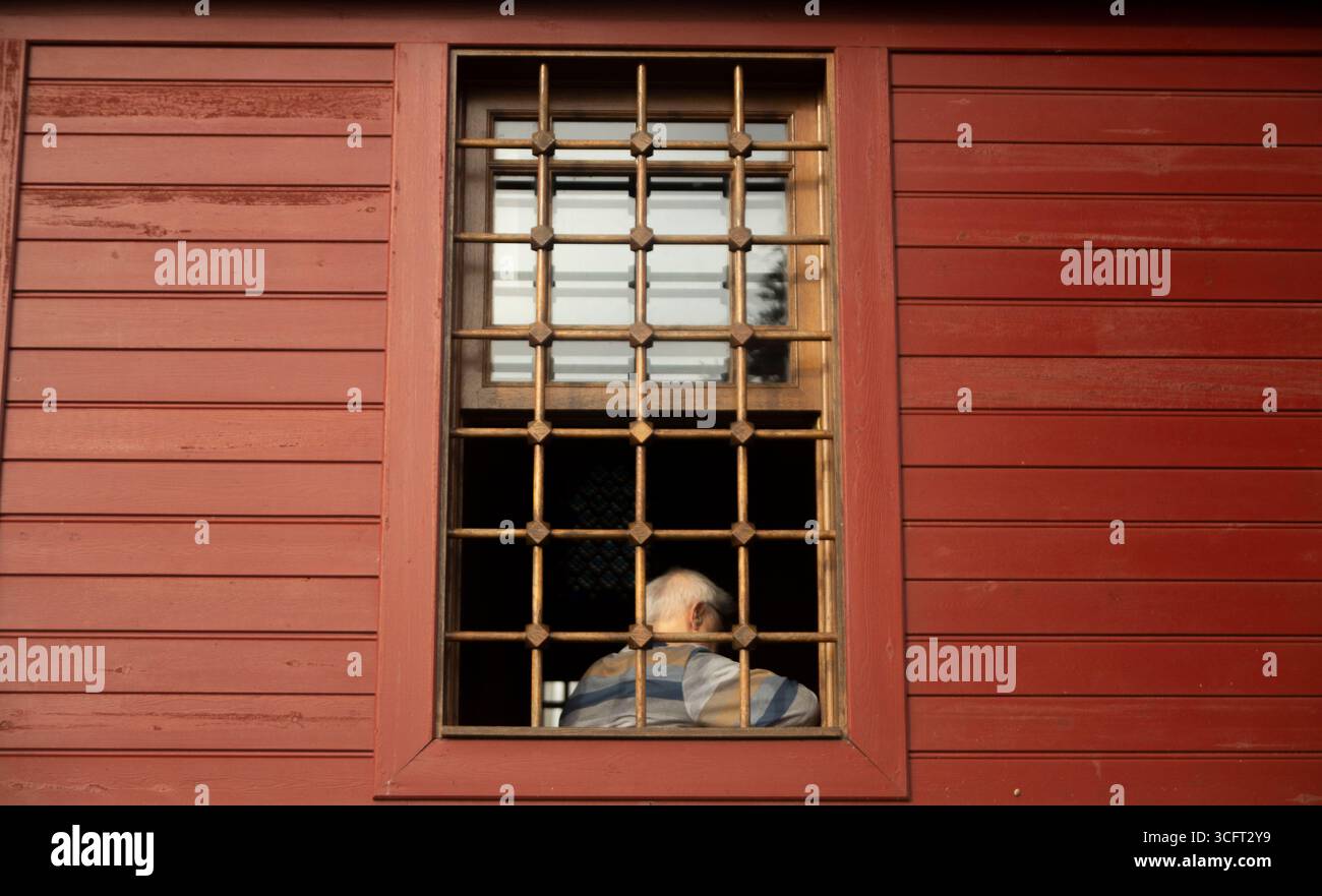 Rückansicht eines älteren Mannes, der durch ein Fenster mit Sicherheitsbügeln blickt Stockfoto
