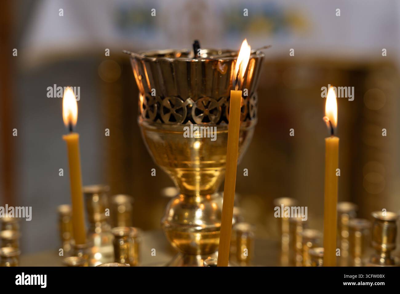 Flackernde Kerzen umgeben eine glänzende Messinglampe, während sich Gläubige in einem ruhigen Raum versammeln, um ein heiliges Ritual zu beobachten. Die Atmosphäre ist erfüllt mit Rev Stockfoto