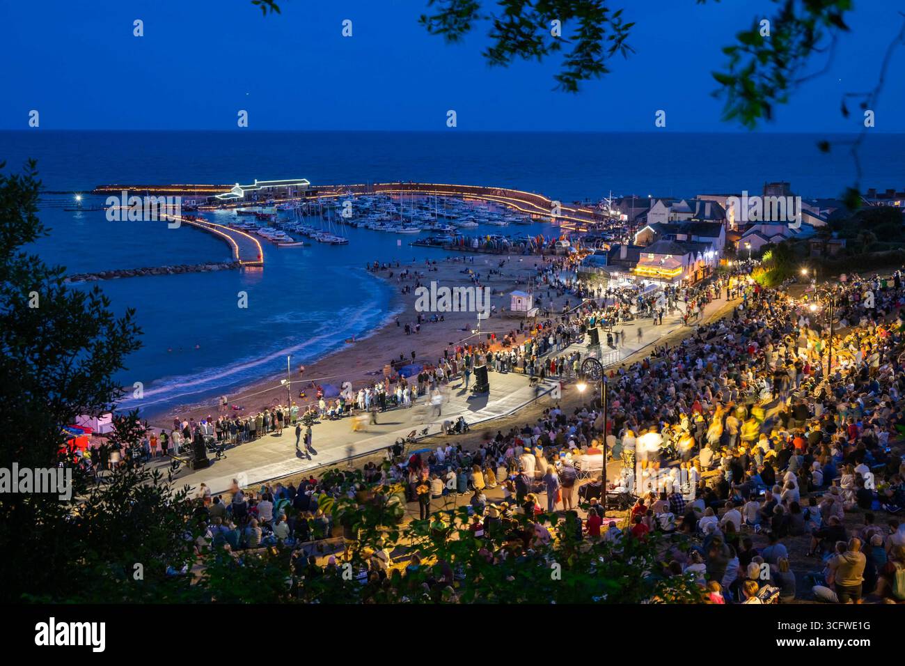 Lyme Regis, Dorset, Großbritannien. August 2025. Tausende von Zuschauern versammelten sich in der Abenddämmerung im Lister Garden und am Meer, um die Candles on the Cobb Illuminations im Badeort Lyme Regis in Dorset zu beobachten. Rund 5000 Kerzen wurden entlang der historischen Hafenmauern von Cobb zum Gedenken an ihre Lieben angezündet. Das gesammelte Geld wird aufgeteilt auf lokale gute Zwecke, Prostate Cancer UK und Macmillan Cancer Support. Bildnachweis: Graham Hunt/Alamy Live News Stockfoto