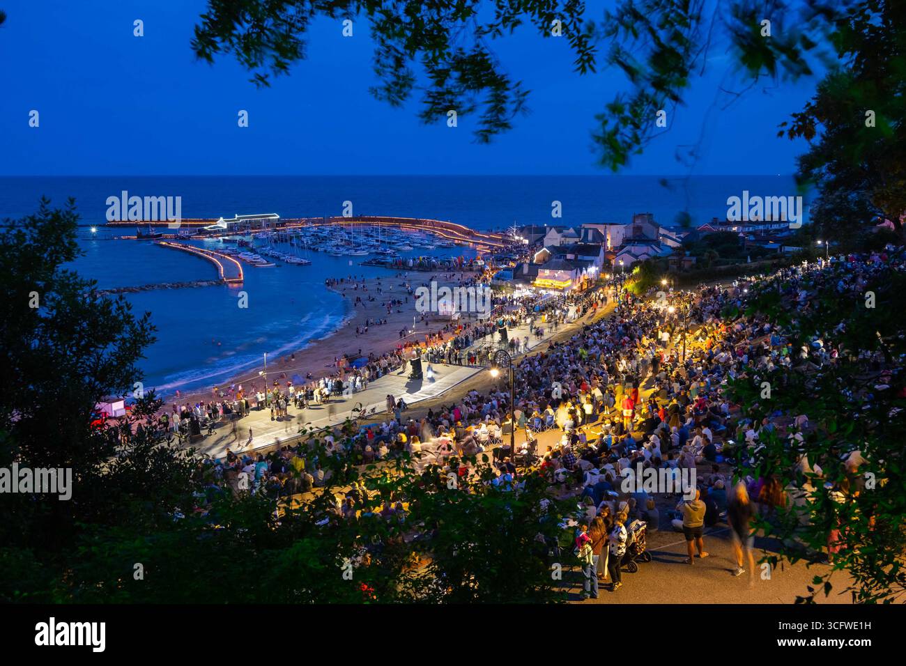 Lyme Regis, Dorset, Großbritannien. August 2025. Tausende von Zuschauern versammelten sich in der Abenddämmerung im Lister Garden und am Meer, um die Candles on the Cobb Illuminations im Badeort Lyme Regis in Dorset zu beobachten. Rund 5000 Kerzen wurden entlang der historischen Hafenmauern von Cobb zum Gedenken an ihre Lieben angezündet. Das gesammelte Geld wird aufgeteilt auf lokale gute Zwecke, Prostate Cancer UK und Macmillan Cancer Support. Bildnachweis: Graham Hunt/Alamy Live News Stockfoto