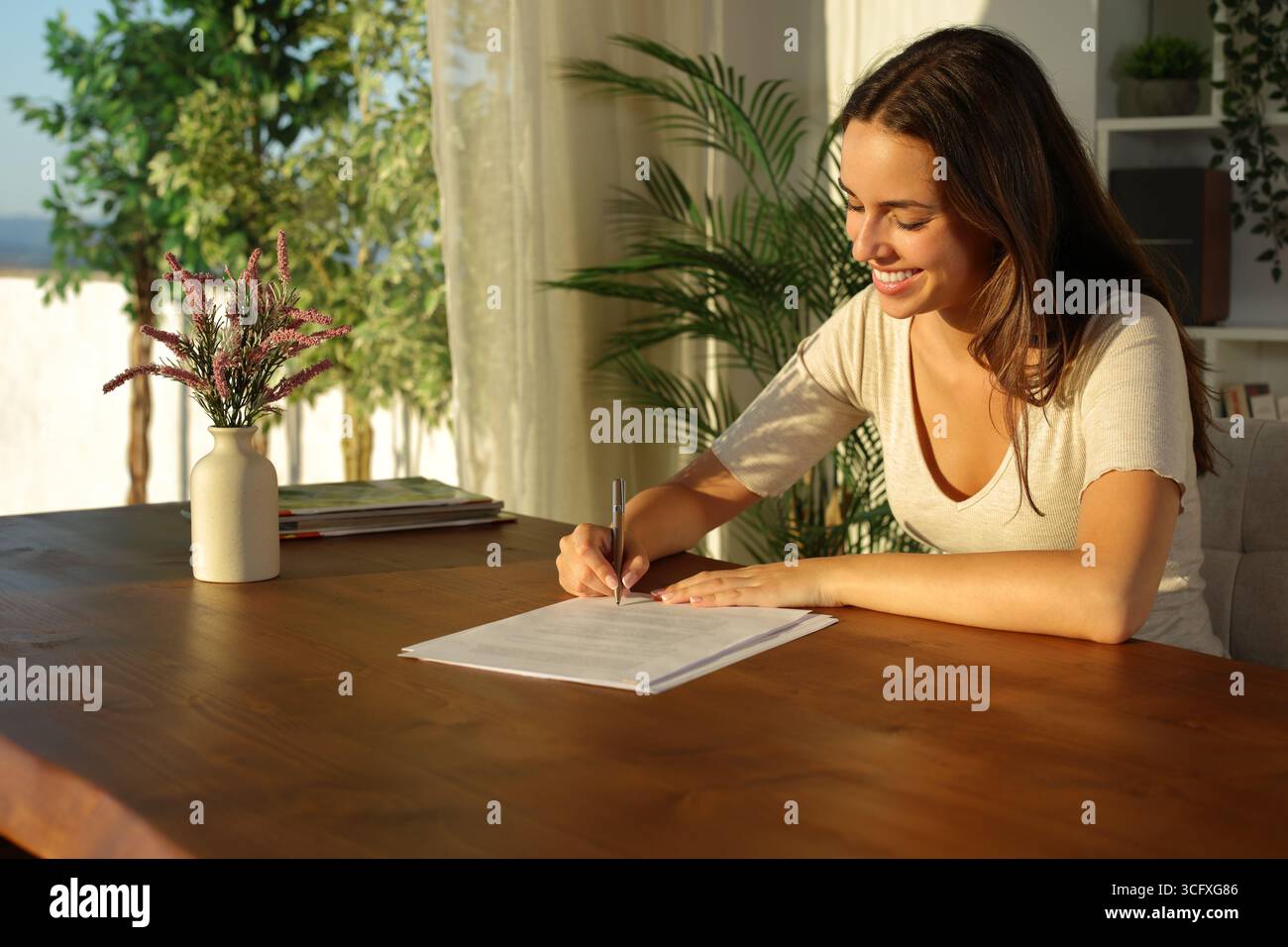 Glückliche Frau, die Dokumente auf Holztisch bei Sonnenuntergang zu Hause unterschreibt Stockfoto