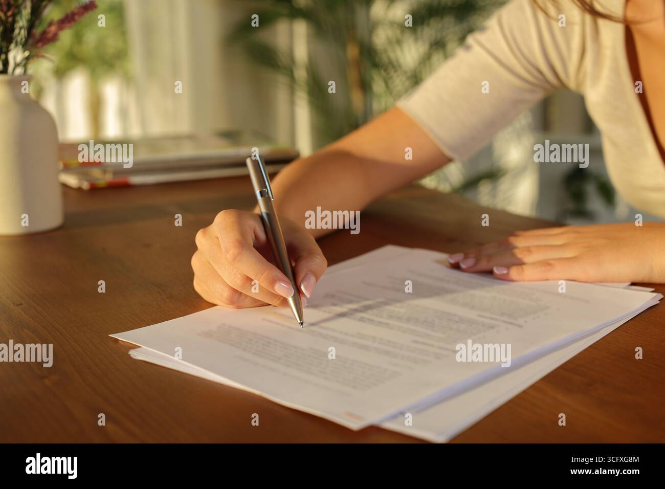 Nahaufnahme einer Frau, die bei Sonnenuntergang zu Hause ein Dokument auf Holztisch ausfüllt Stockfoto