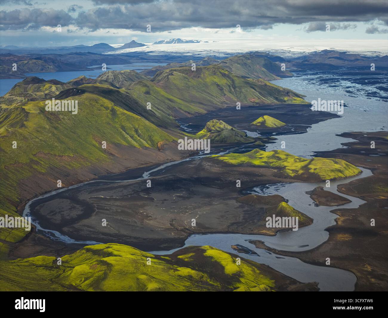 Blick aus der Vogelperspektive auf das grüne Hochland, das auf die geflochtenen Kanäle des Gletscherflusses trifft, im Kontrast zu dunklem vulkanischem Sand unter einem riesigen isländischen Himmel, Hochland, Island. Stockfoto