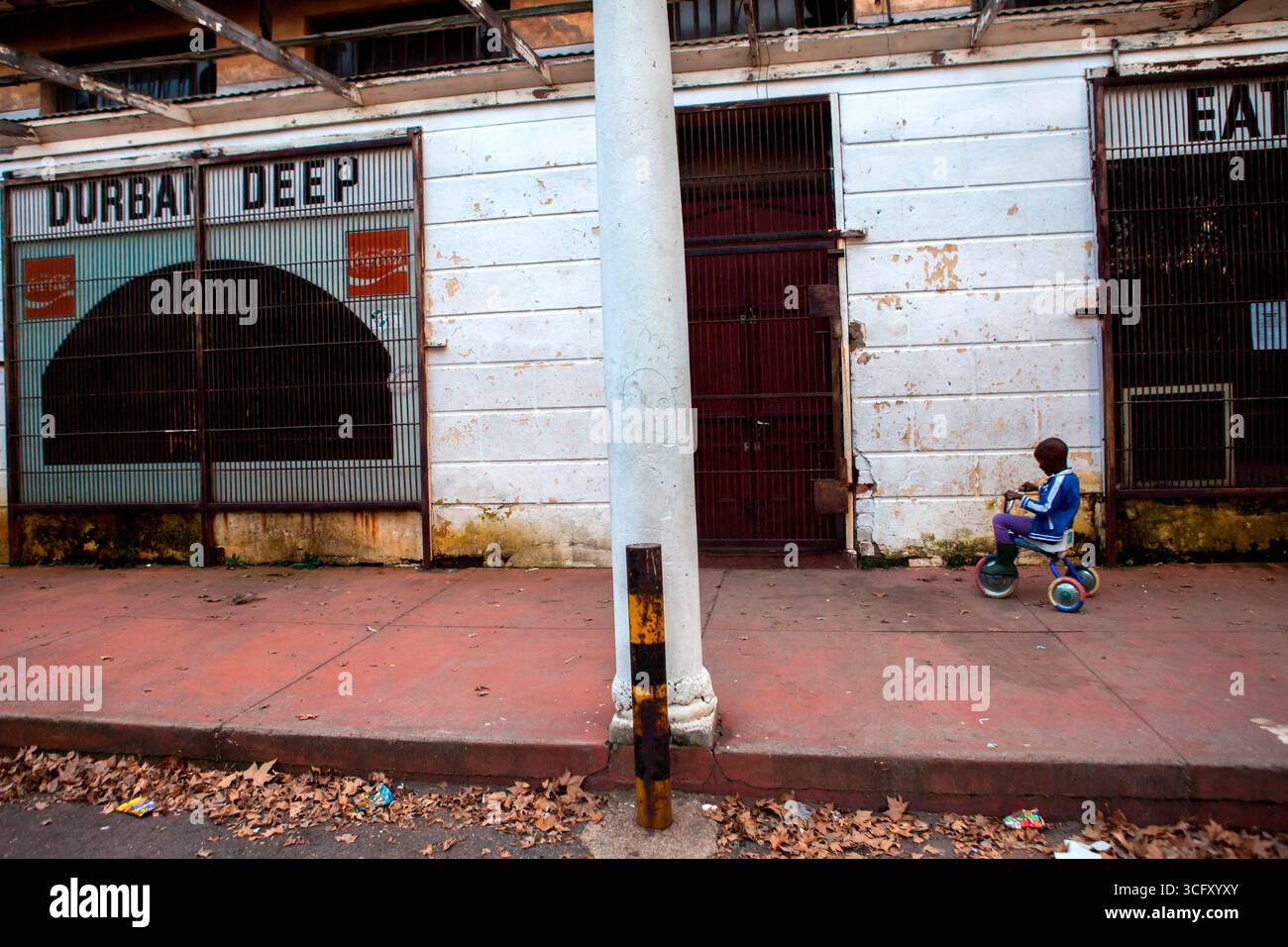 Ein Kind fährt ein Dreirad vor einem inzwischen geschlossenen Restaurant neben einem der alten Hostels von Durban Deep Goldmine. Stockfoto