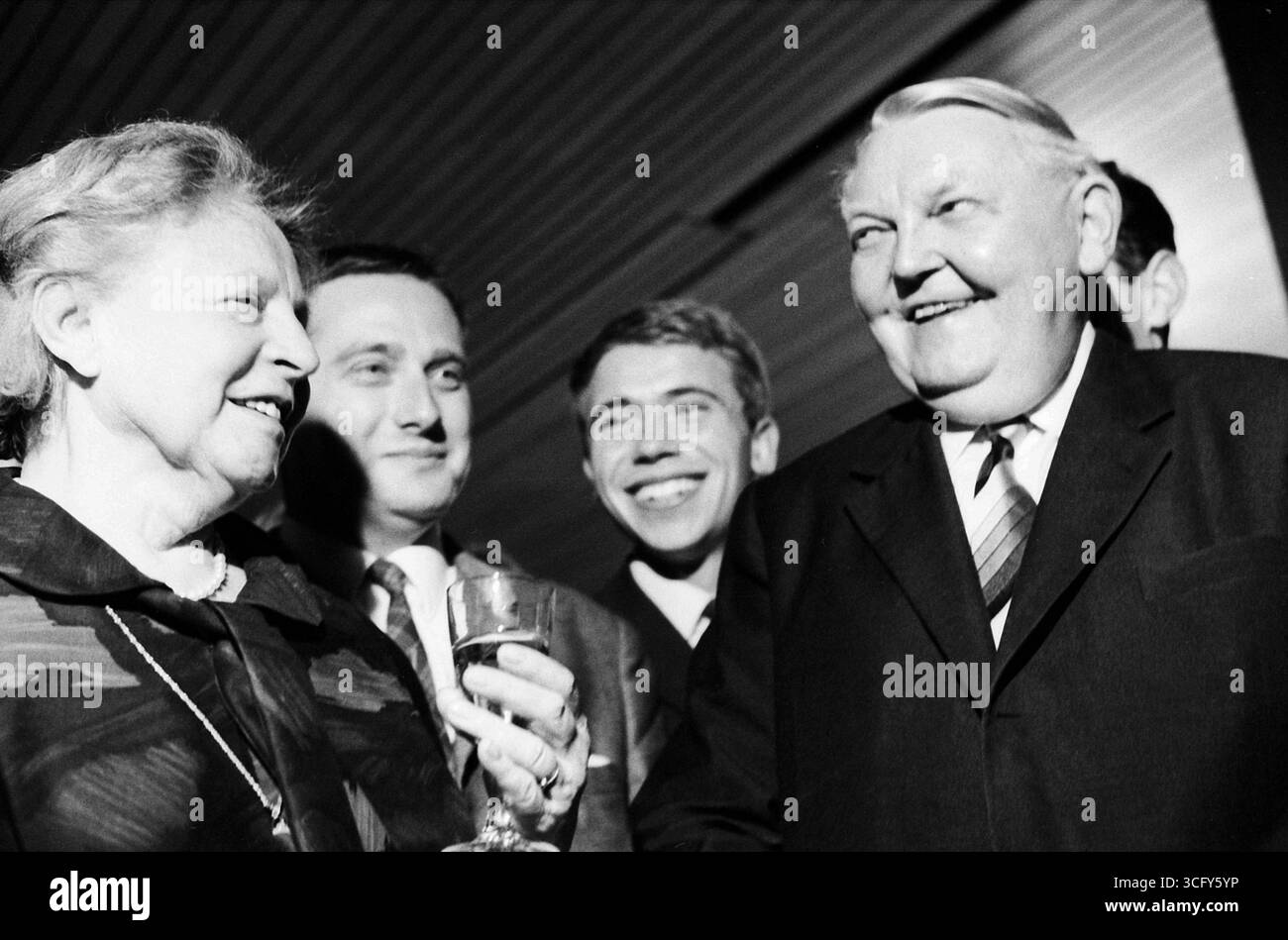 Ludwig Erhard und seine Ehefrau Luise Erhard stehen gemeinsam mit mehreren jüngeren Männern bei einem festlichen Anlass. Beide lachen, Luise Erhard hält ein Glas in der Hand. Stockfoto