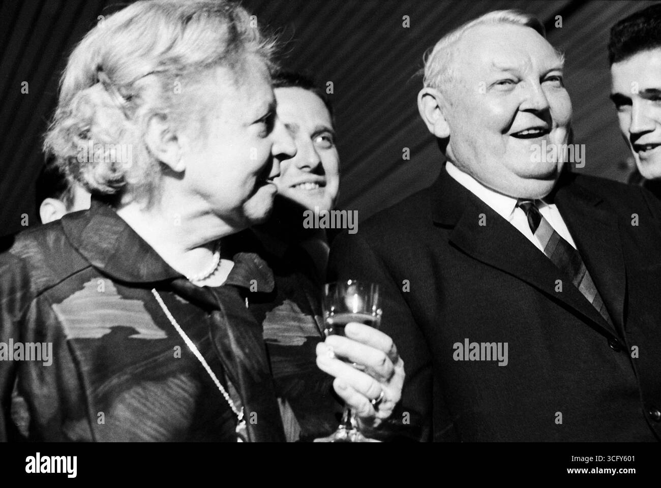 Ludwig Erhard und seine Ehefrau Luise Erhard stehen gemeinsam mit mehreren jüngeren Männern bei einem festlichen Anlass. Beide lachen, Luise Erhard hält ein Glas in der Hand. Stockfoto