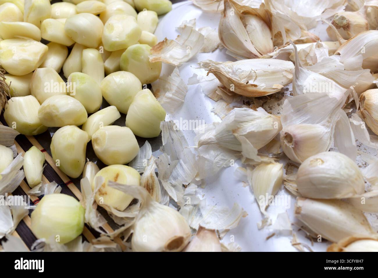 Frische Knoblauchzehen werden geschält und neben ihren Schalen auf eine weiße Oberfläche gelegt, um einen kulinarischen Zubereitungsprozess zu ermöglichen. Stockfoto