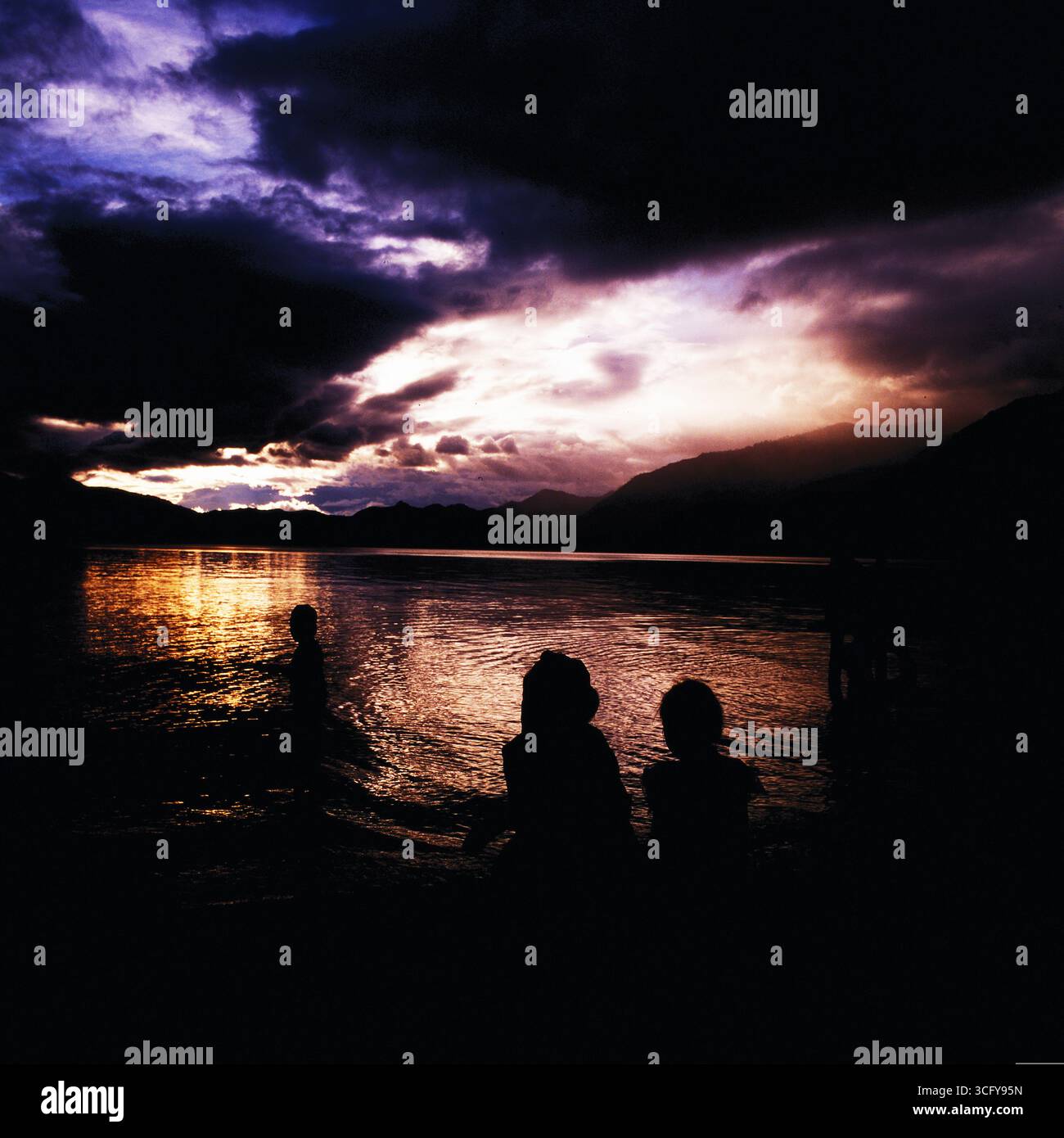 Silhouetten von Menschen am Ufer eines Sees bei Sonnenuntergang, umgeben von Bergen und dramatischem Wolkenhimmel. Guatemala, um 1980. Stockfoto