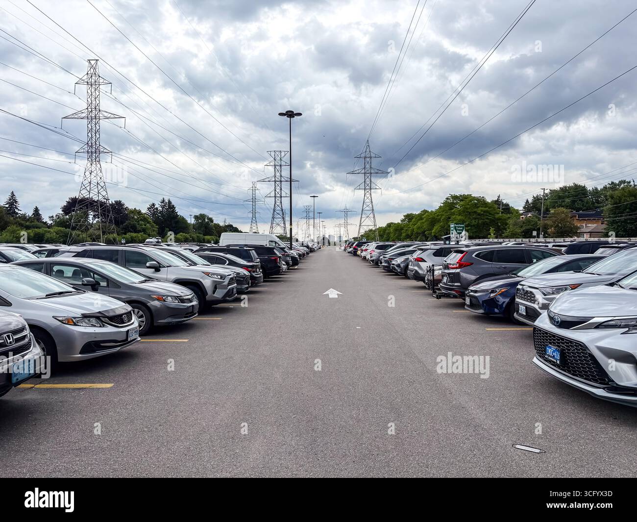 Ein großer Außenparkplatz voller Reihen geparkter Autos unter einem bewölkten Himmel. Markante Strommasten und Freileitungen erstrecken sich über Acros Stockfoto