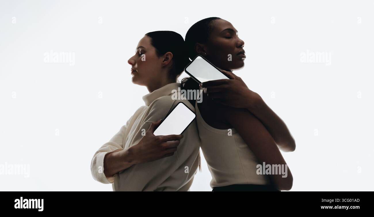 Silhouetten von zwei Frauen, die Rücken an Rücken stehen und Smartphones mit leeren Bildschirmen halten, isoliert auf weißem Hintergrund. Perfekt für Mockups von mobilen Bildschirmen Stockfoto