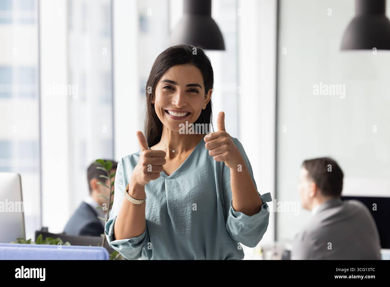 Geschäftsfrau, die im Büro steht, in die Kamera schaut, Daumen nach oben zeigt Stockfoto