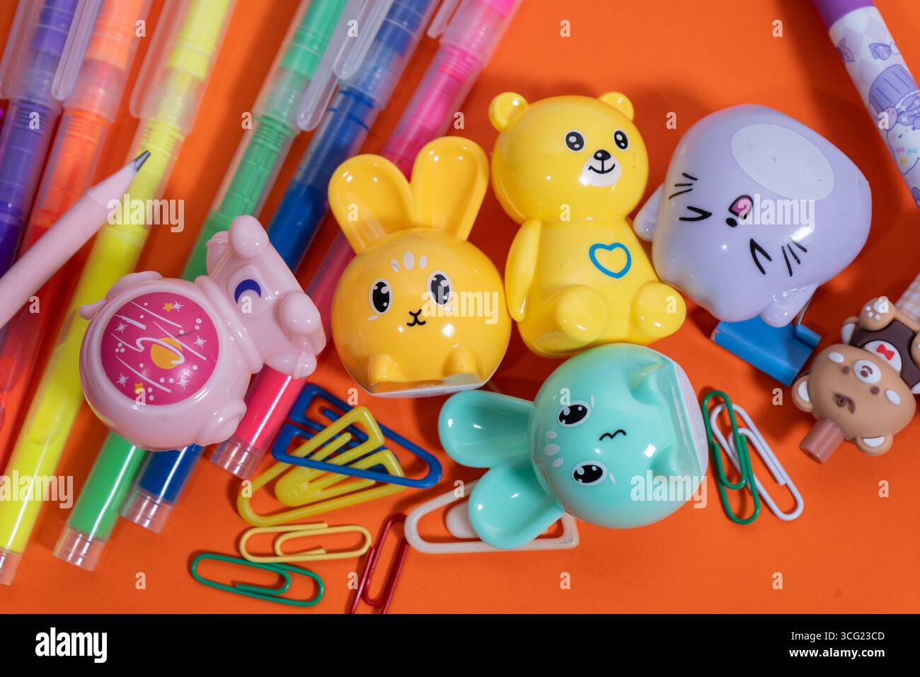 Niedliche tierförmige Bleistiftspitzer, bunte Stifte und Büroklammern schaffen ein lebendiges Arrangement auf orangem Hintergrund Stockfoto