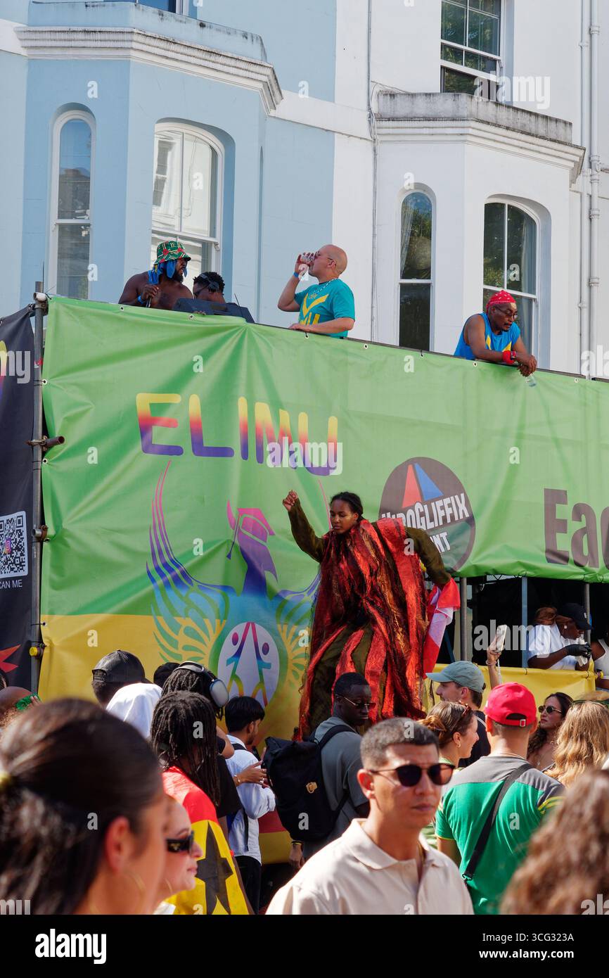 Frau auf Stelzen in rotem Mantel neben einem Wagen inmitten der Menschenmassen während des Notting Hill Carnival in London. England, 25. August 2025 Stockfoto