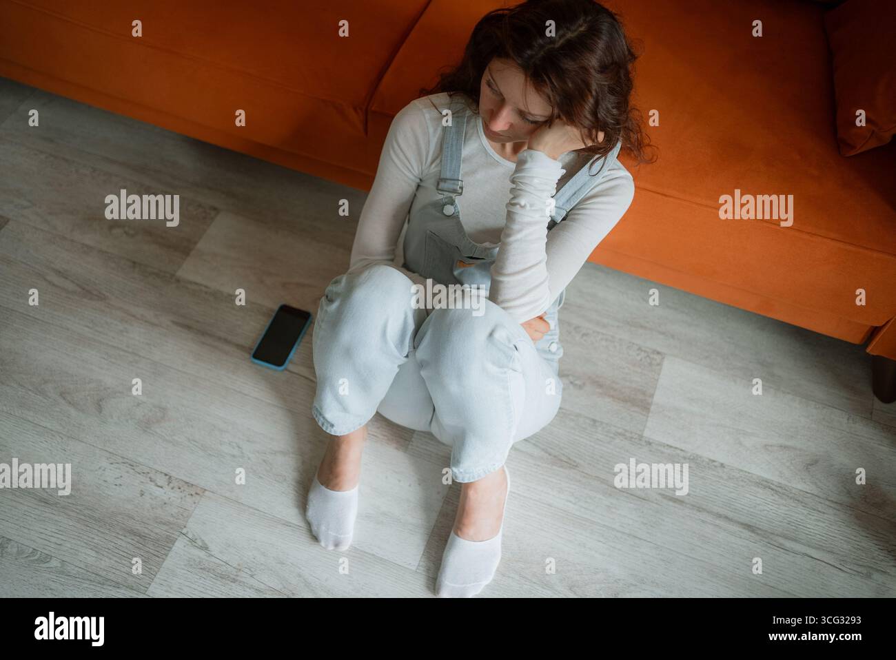 Traurige Frau, die neben einem orangen Sofa auf dem Boden sitzt, Smartphone in der Nähe, und sich von Einsamkeit und Verzweiflung überwältigt fühlt, nachdem sie die harten Realitäten des Cybermobbing erlebt hat Stockfoto