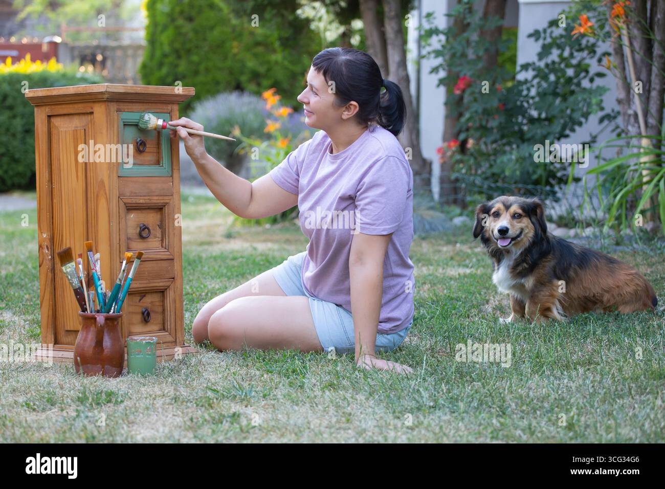 Frau, die Holzmöbel im Hof mit ihrem Hund malte. Die Innendekoration wird neu gestaltet Stockfoto