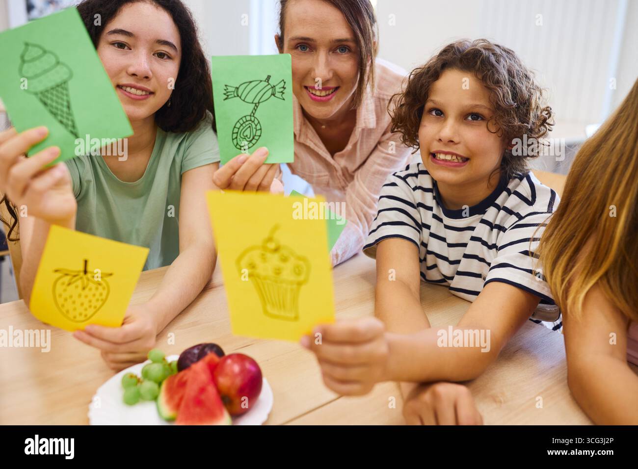 Eine Gruppe von Kindern und ihrem Lehrer zeigen handgezeichnete Essenskarten während einer Lernaktivität. Frisches Obst schmückt den Tisch und schafft so eine lustige lea Stockfoto