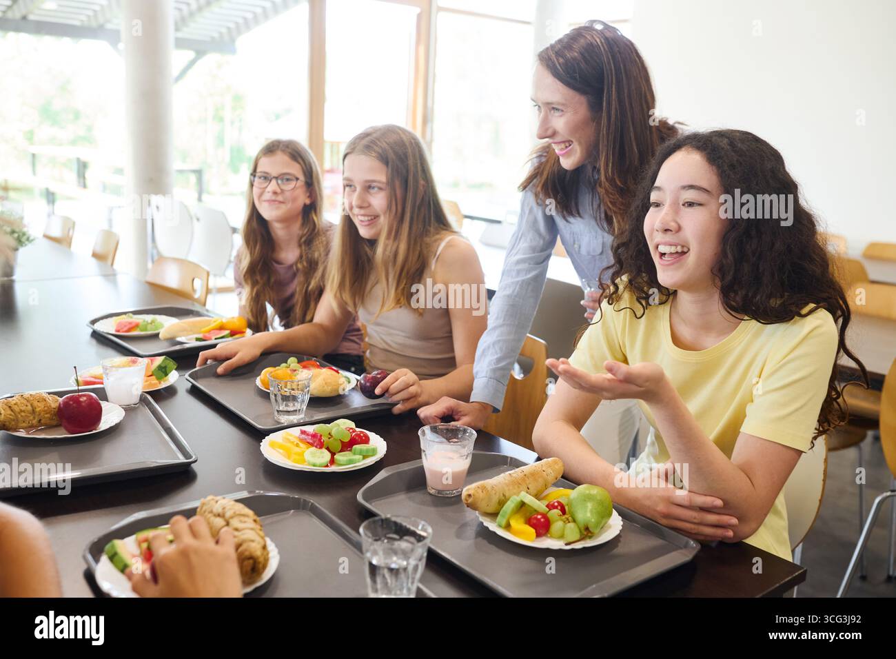 Eine Gruppe von Schülern lacht, während sie frische und nahrhafte Mahlzeiten in einer gut organisierten Schulcafeteria genießen, um Kameradschaft und Freude zu fördern. A te Stockfoto