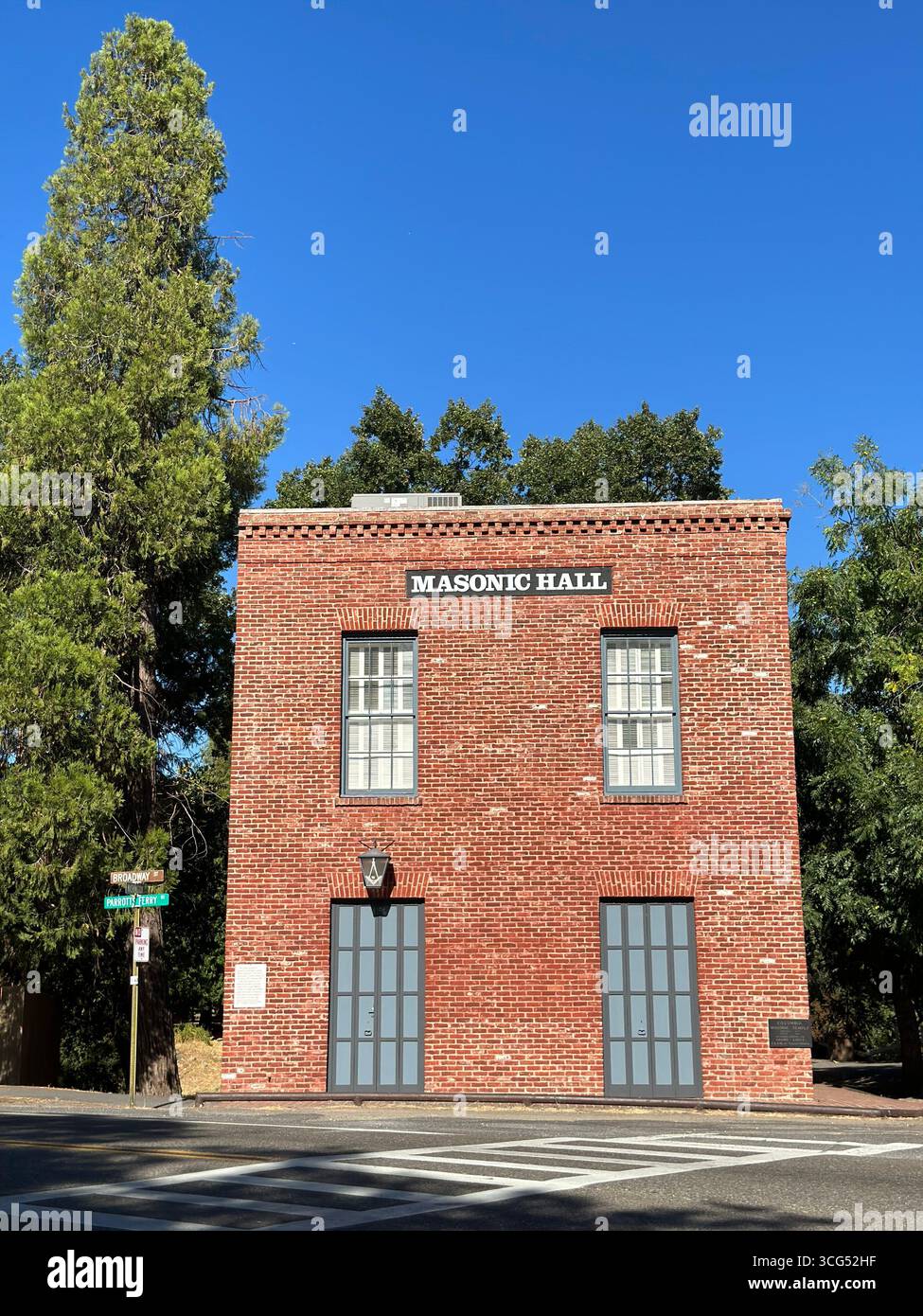 An der Ecke Broadway und Parrott Street in Kalifornien steht eine historische Freimaurerhalle aus roten Backsteinen, die von üppigen grünen Bäumen eingerahmt ist. - Smartphone-aufgenommenes Stockfoto