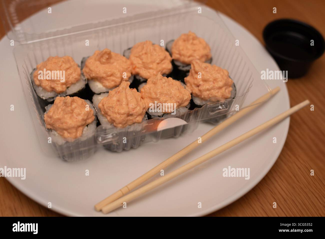 Köstliche Sushi-Brötchen mit einer cremigen, scharfen Sauce in einer durchsichtigen Verpackung, serviert mit Essstäbchen auf einem weißen Teller auf einem Holzteller Stockfoto