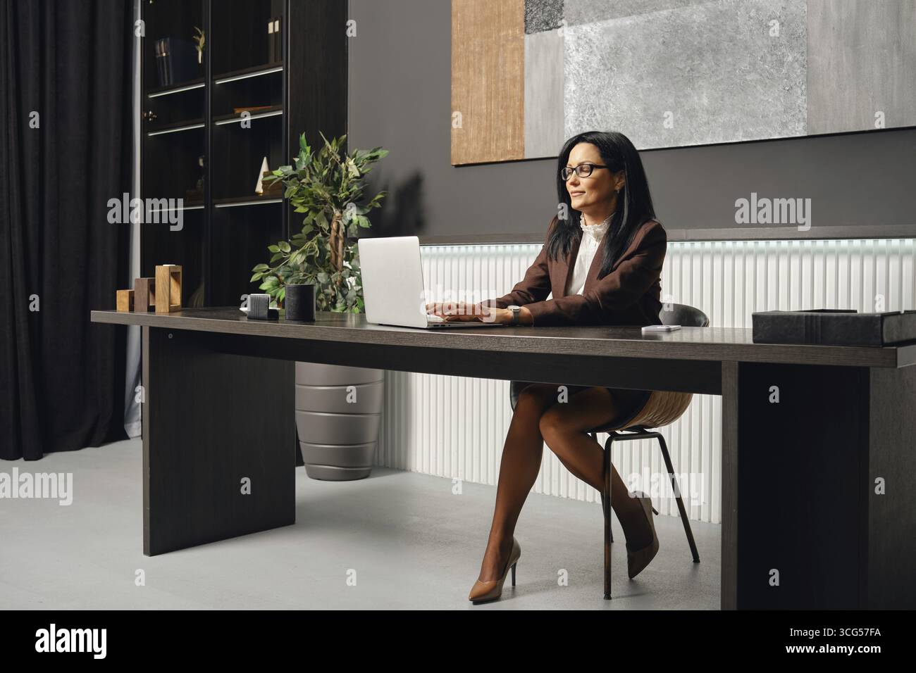 Eine Geschäftsfrau, die einen Bericht auf ihrem Laptop schreibt, während sie an einem eleganten Schreibtisch in einem modernen Büro sitzt Stockfoto