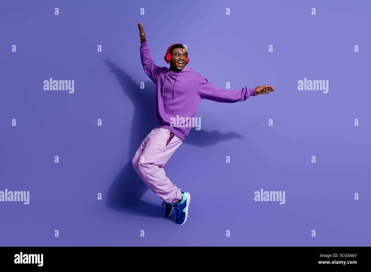 Energiegeladener junger Mann in leuchtendem lila Outfit, lächelt und tanzt mit dynamischer Pose vor farbigem Studiohintergrund Stockfoto