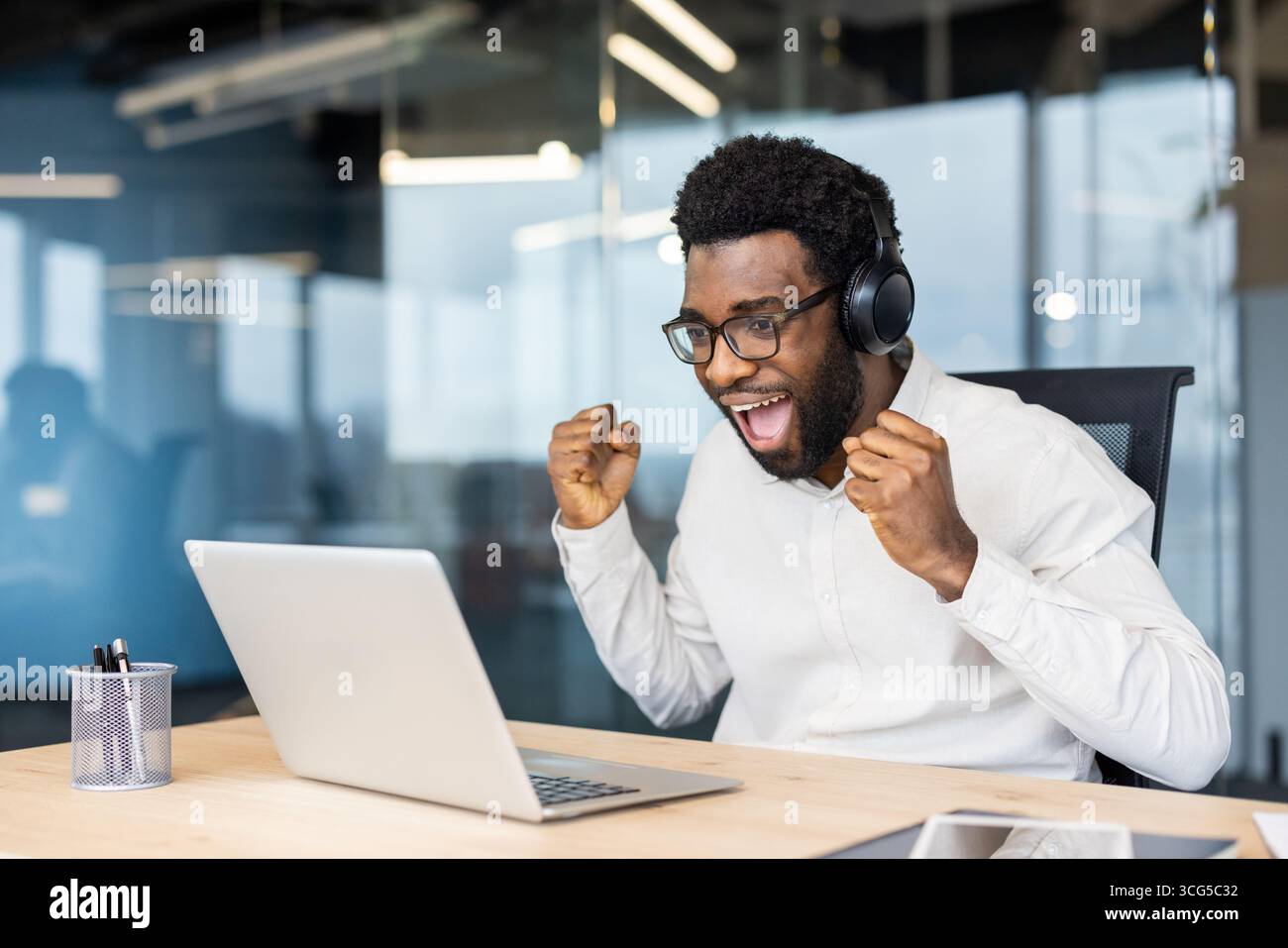 Ein fröhlicher Mann mit Kopfhörern feiert mit Begeisterung den Erfolg in einem modernen Büroarbeitsplatz. Sein Laptop ist auf dem Schreibtisch sichtbar und zeigt eine lebendige und inspirierende Büroumgebung. Stockfoto