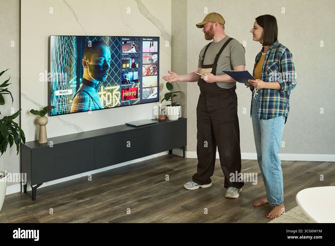 Kaukasischer Mann mittleren Alters mit Mütze, der neben kaukasischer junger erwachsener Frau steht, die ein Klemmbrett hält, und beide diskutieren über Streaming-Dienste, die auf großen Flachbildfernsehern gezeigt werden Stockfoto