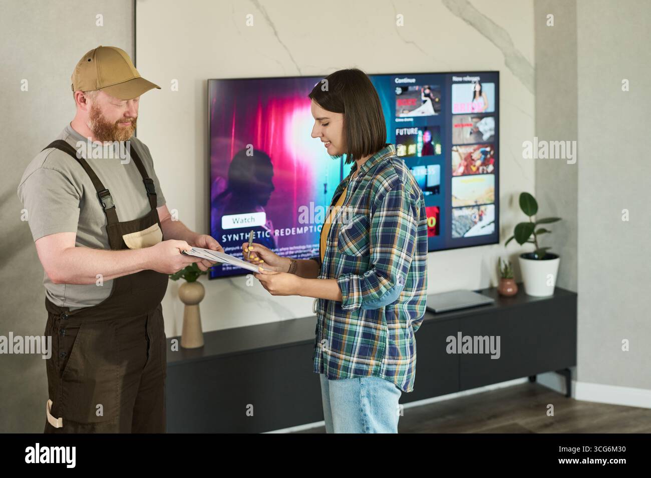 Kaukasischer Mann mittleren Alters, der Uniform trägt und Dokumente an junge Erwachsene asiatische Frau übergibt, die in einem modernen Wohnzimmer ein Klemmbrett unterzeichnet Stockfoto