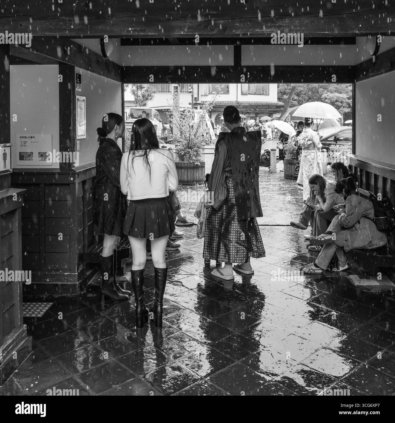 Ein Schwarzweiß-Bild, das eine Gruppe von Menschen darstellt, die unter einer Struktur im japanischen Stil Schutz vor dem Regen suchen, die kulturelle Kleidung und Mod widerspiegelt Stockfoto