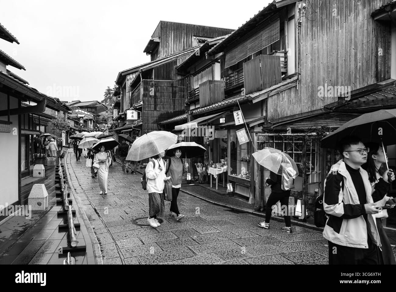 Schwarzweiß-Foto von Menschen, die bei regnerischem Wetter in einer historischen Straße spazieren gehen. Zeigt traditionelle Architektur, wobei Einzelpersonen um halten Stockfoto